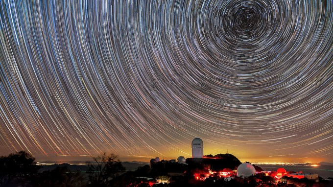 Stars swirl across the sky, creating a circular pattern in this time-lapse photo. In the foreground on a small rise of land is the Mayall Telescope at Kitt Peak National Observatory in Arizona, which is conducting the DESI dark energy survey.