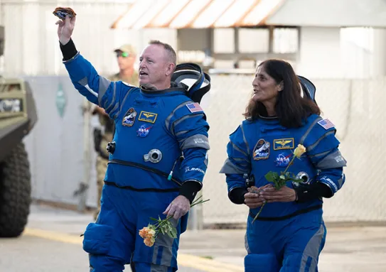 Butch Wilmore and Suni Williams walking outside while wearing blue Boeing space suits.