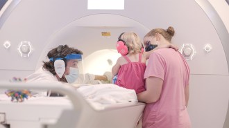 A baby wearing pink headphones gets ready to go into a functional MRI machine for a memory test. Two helper adults are nearby.