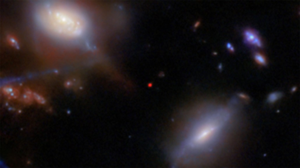 Several blurry galaxies of various sizes and shapes on a black background in this image from the JWST. In the center, a small red dot marks the most distant galaxy in this field, JADES-GS-z13-1.