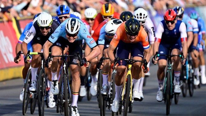 A group of professional cyclists in a tight sprint finish during a race. The riders are wearing colorful team uniforms and helmets, gripping the handlebars with intense focus. Crowds are in the background.