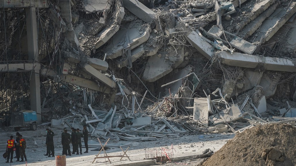 A photo of a building in Bangkok crumbled by a magnitude 7.7 earthquake in Malaysia on March 28, 2025. Onlookers stand in the shadow of the large gray concrete slabs from the crushed building.