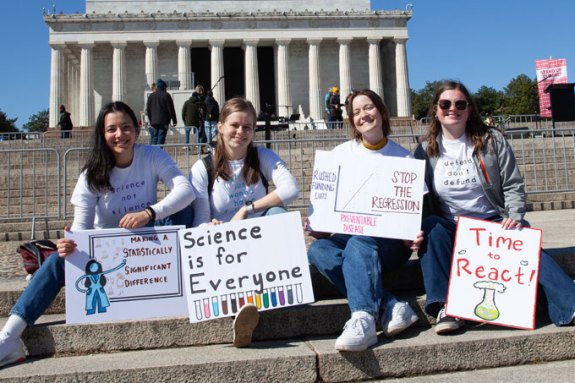 Four young women hold signs reading "Making a statistically significant difference," "Science is for everyone," "Stop the regression" and "Time to react."