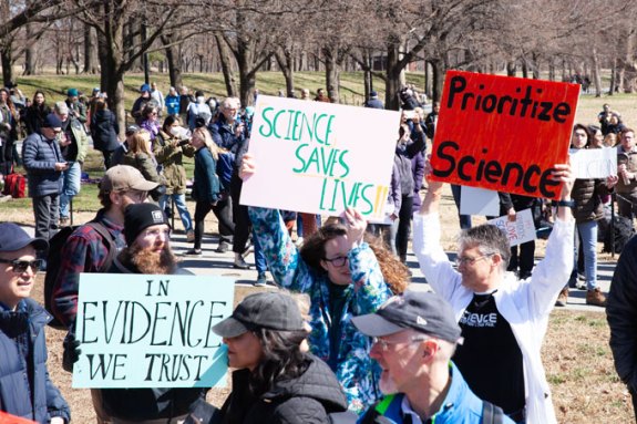 People hold signs reading "In evidence we trust," "Science saves lives" and "Prioritize science."