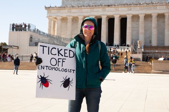 A young woman in front of the Lincoln Memorial holds a sign reading TIcked Off Entomologist.