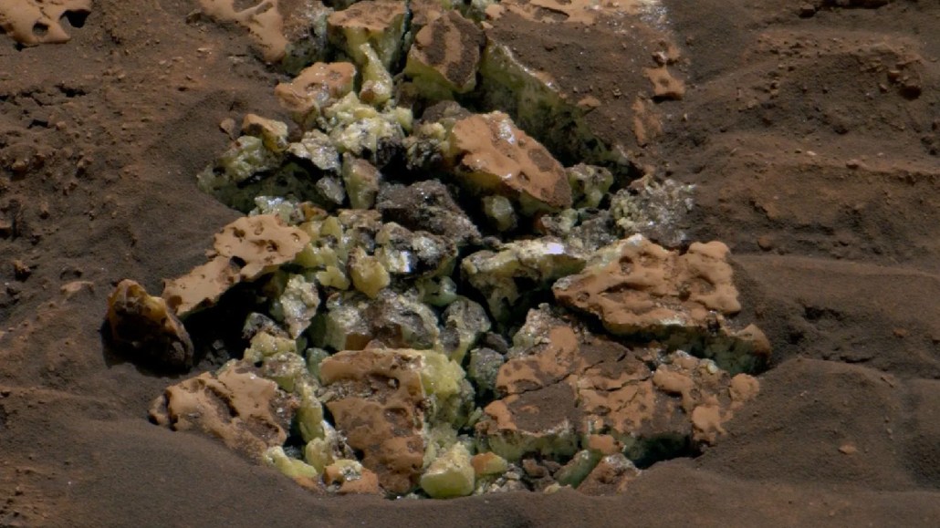 A close-up view of reddish dirt, with yellow crystals emerging from broken rocks in the middle