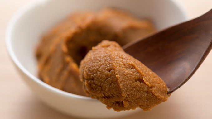 A wooden spoon with nutty brown miso paste in the foreground and a white bowl with miso in the background.