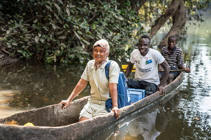 Three people sit in a wooden boat floating on a river. A blue cooler sits between the first person. They are delivering vaccines to people in remote locations in Sierra Leone.