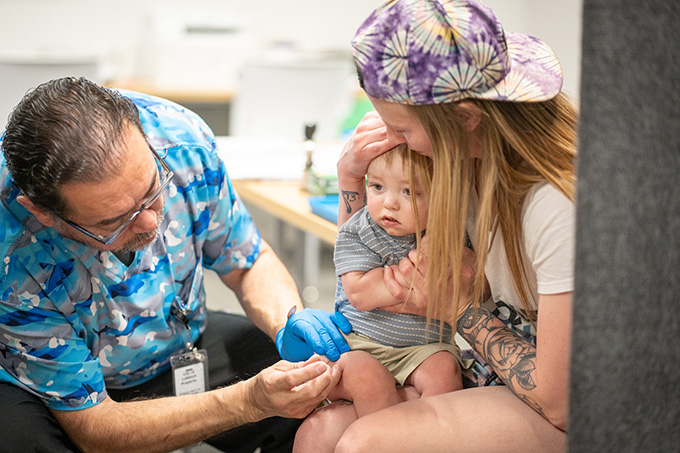 A health-care worker prepares to give a young child a measles vaccine the thigh. The boy sits on his mother's lap, which she holds his hands and cradles his head.