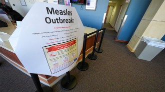 A large white sign outside a hospital corridor proclaims in large black letters, Measles Outbreak. The sign gives symptoms of the disease and has a graphic with information about how to protect children with measles vaccines. A second child in Texas recently died from measles.