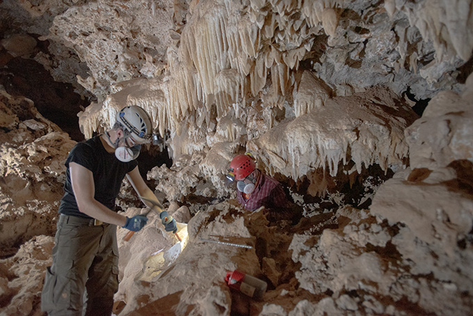 Two researchers wearing helmets use tools to take samples from inside a cave. The interior is filled with rock formations like stalactites hanging from the cave ceiling.