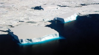 An aerial view of flat-topped icebergs floating in the dark waters off the coast of Antarctica, with fragmented sea ice covering the surrounding area.