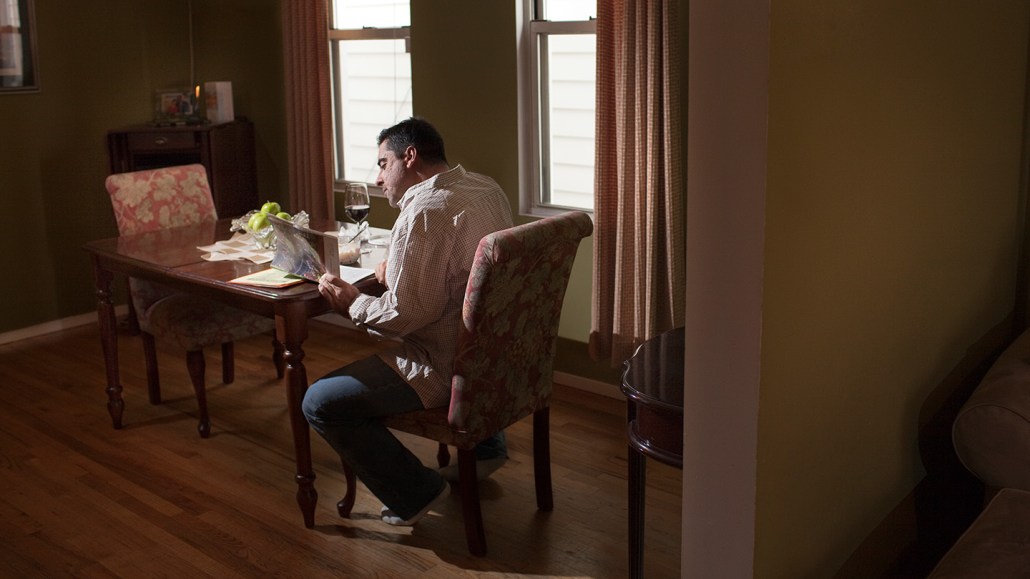 A man sitting alone at a dining table in a dimly lit room, reading something, with a glass of red wine and a partially eaten meal in front of him.