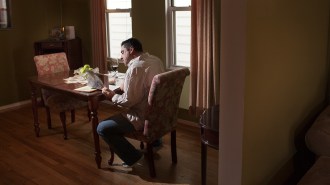 A man sitting alone at a dining table in a dimly lit room, reading something, with a glass of red wine and a partially eaten meal in front of him.