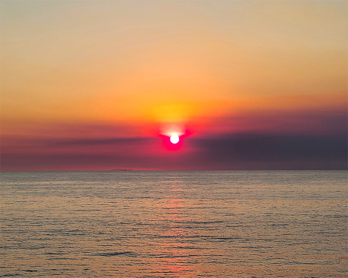 The sun slightly above the waterline of the ocean. Cloudy smoke makes the setting sun appear hazy.