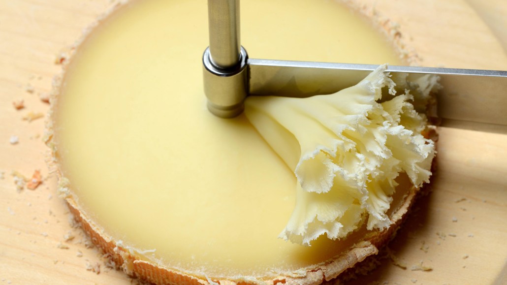 A utensil called a girolle with a wheel of cheese, likely tête de moine, with the post of the girolle going through its center and a blade that rotates around the post, which has scraped off a frilly rosette