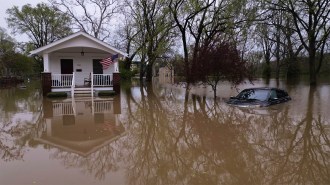 Muddy-looking water surrounds a car and house, creeping up toward the car windows and the steps of the front porch.