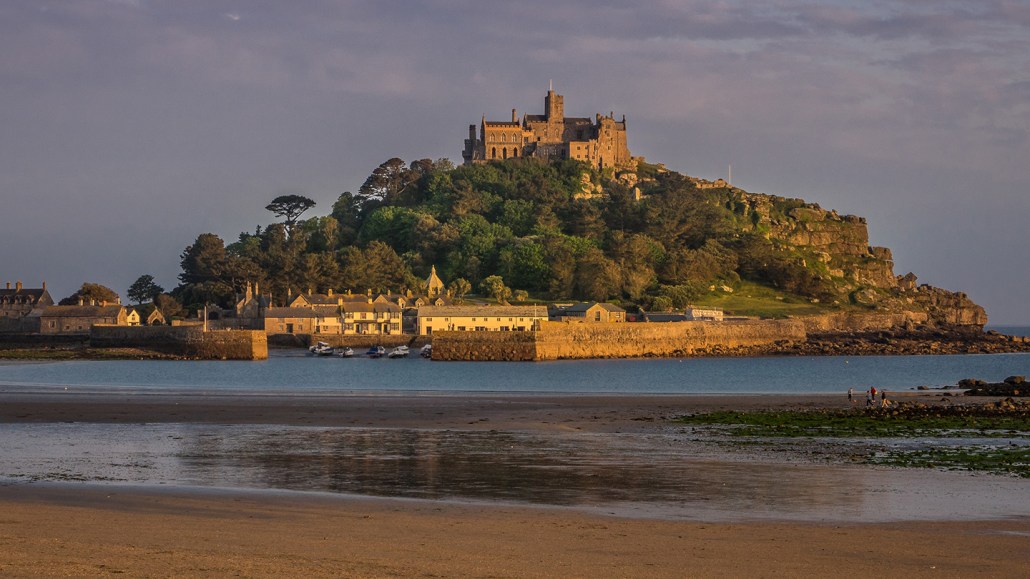 The small British island of Saint Michael's Mount pictured from a nearby beach, with trees and buildings dotting the small island in golden light
