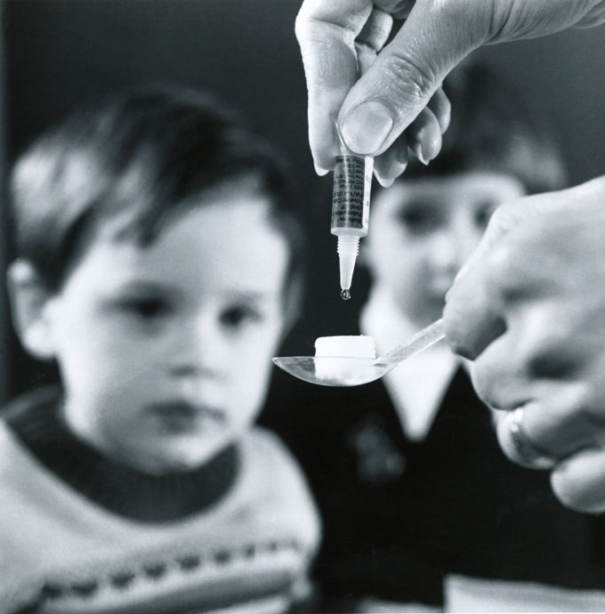 In the foreground, a person's hand squeezes a tube so that liquid drops onto a sugar cube being held in a spoon by their other hand. In the background are two children staring at the sugar cube.