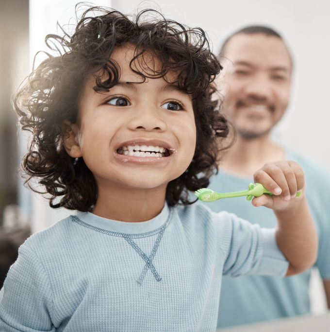 A young child with dark, curly hair and big, brown eyes wearing a light blue knit top and holding a green toothbrush shows their teeth. Flecks of toothpaste foam dot their lips. A smiling man with short dark hair, beard and mustache and a blue shirt who is blurred in the background looks on.