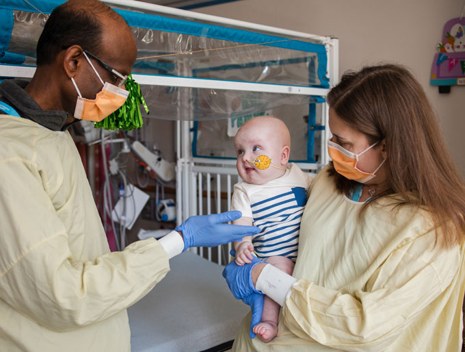 Two doctors interact with baby for whom they designed a gene therapy. On the left a doctor wearing a yellow gown, orange surgical mask, and glasses reaches out blue-gloved hands toward the baby who has a big bird sticker on his cheek holding a feeding tube in his nose. Another doctor with long brownish hair also wearing a yellow robe and orange surgical mask holds the baby who is wearing a white outfit with horizontal blue stripes.
