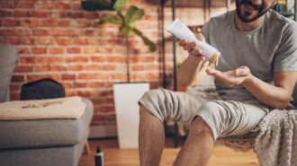 A man sitting in a chair with his pants legs rolled up to his knees squeezes lotion onto his hand, as if preparing to moisturize his skin.