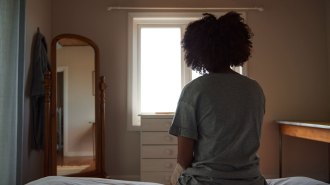 A woman with her back to the camera sits on a bed in a dimly lit room looking out the window