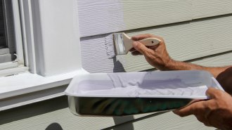 A close-up from the elbows down of a person painting the siding of a house. One hand holds a paintbrush while the other holds a tray of white paint