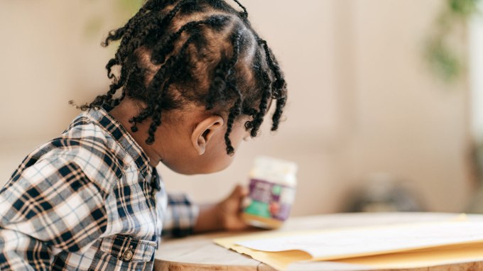 A young toddler wearing a flannel shirt sits at a table and holds a jar of food.