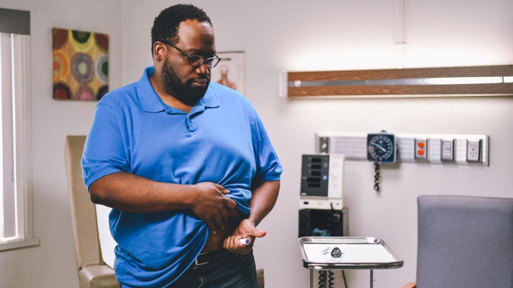 A man injects himself in the stomach with a "pen" containing a GLP-1 drug. The man has dark skin and hair, is wearing glasses and wearing a blue polo-style shirt. He is lifting his shirt to expose the spot on his stomach where he will inject the medication. He appears to be in a doctor's office or hospital room.