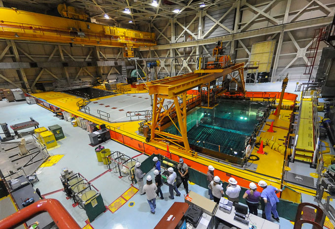 People in hard hats walk past a water pool containing spent nuclear fuel.