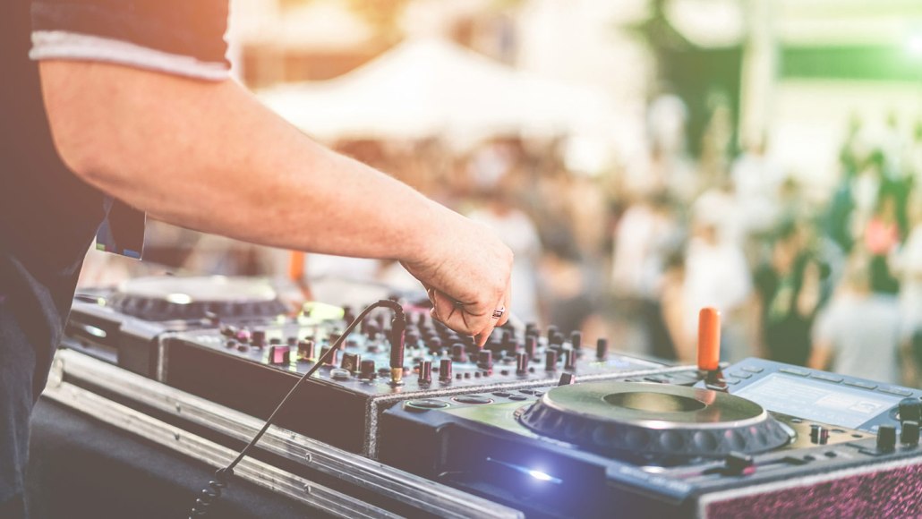 A close-up of hands at a DJ table with a blurred summertime crowd in the background