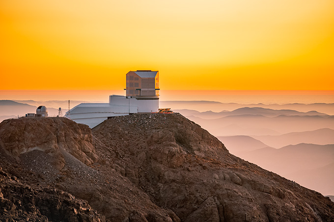 The Rubin Observatory on top of a bald mountain during an orange sunset. Other mountains are visible in the distance.