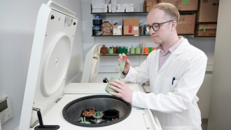 A scientist in a white lab coat operating a centrifuge machine in a laboratory, handling a green rotor with sample tubes, with lab equipment and storage boxes on shelves in the background.