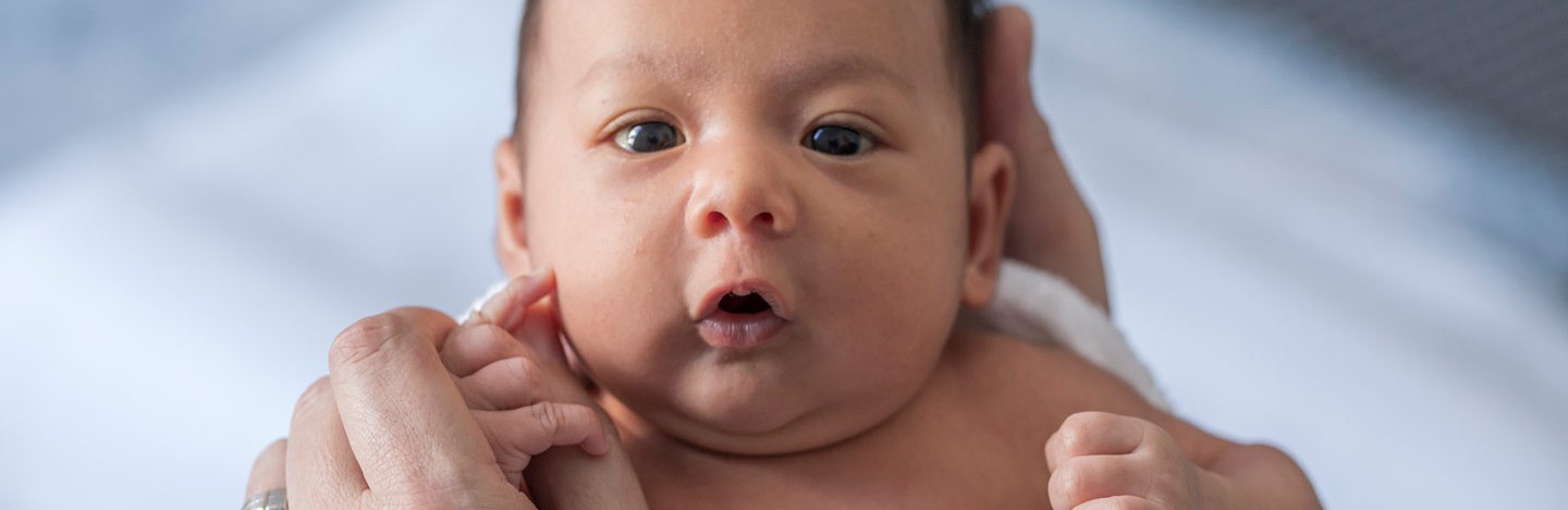 Close up of a one-week-old baby boy with dark hair and eyes. He is wrapped in a white towel and clutching the thumb of an adult wearing a gold ring. His lips are pursed as if cooing.