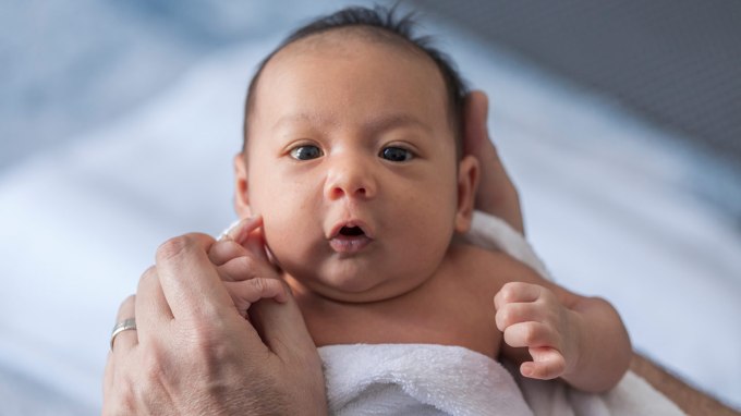 Close up of a one-week-old baby boy with dark hair and eyes. He is wrapped in a white towel and clutching the thumb of an adult wearing a gold ring. His lips are pursed as if cooing.