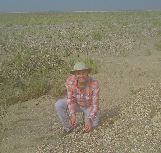 A researcher in jeans, a plaid long-sleeve shirt and sunhat kneels on sandy, rocky soil, holding tools to search the ground for fossils.