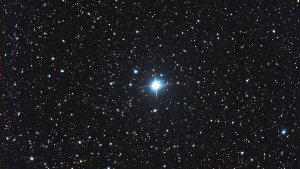 The exploding star V572 Velorum appears like a blue glowing orb with four rays extending out in a cross shape. It sits amid much dimmer blue and yellow stars and an inky night sky.