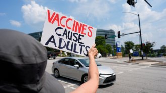 A protestor wearing a black bucket hat holds out a hand-lettered white sign as silver car passes. The sign says "Vaccines cause adults." Vaccines is written in red and cause adults is in black.