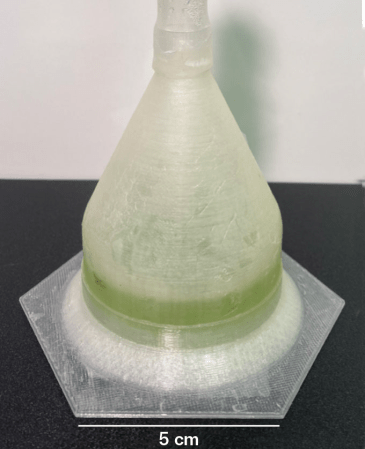 A translucent beaker sits on a black tabletop. the beaker has a hexagon shaped bottom. A small amount of green liquid is inside, where the algae is growing. The liquid fills about a fifth of the container.