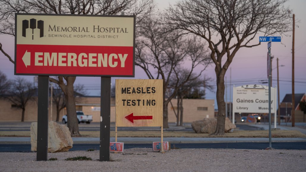 A wooden sign is shown positioned along a road with a spray painted message that says "measles testing" in black. A red arrow points to the left. The sign is next to a permanent sign for Memorial Hospital, Seminole Hospital District.
