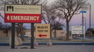 A wooden sign is shown positioned along a road with a spray painted message that says "measles testing" in black. A red arrow points to the left. The sign is next to a permanent sign for Memorial Hospital, Seminole Hospital District.