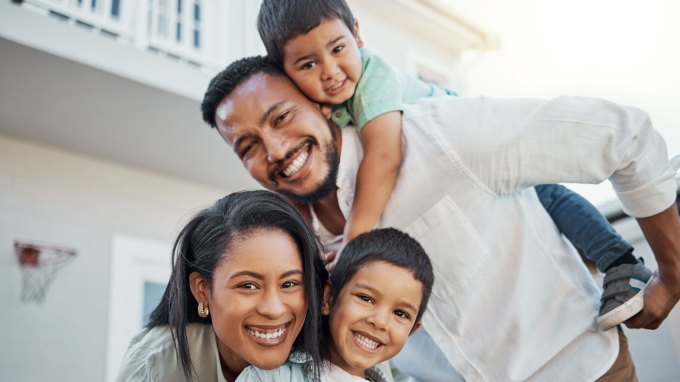 Smiling family of four outdoors, with a man giving a piggyback ride to a young boy, while a woman leans forward beside them, holding another smiling boy in front.