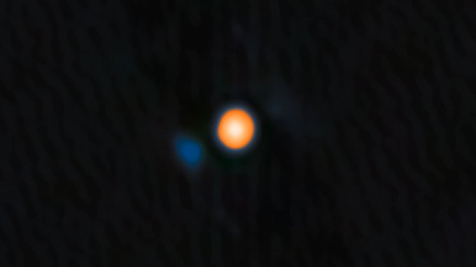 Blurry astronomical image showing the star Betelgeuse as a bright, orange circular blob against a dark background with a smaller, faint blue spot to the lower left.