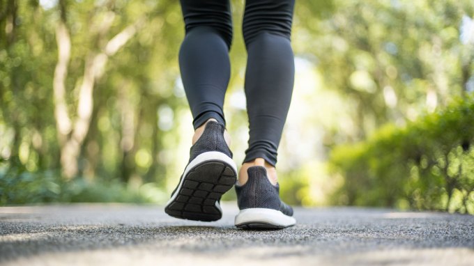 Close-up of a person walking on a paved outdoor path, wearing black leggings and athletic shoes, with green trees and foliage in the background.