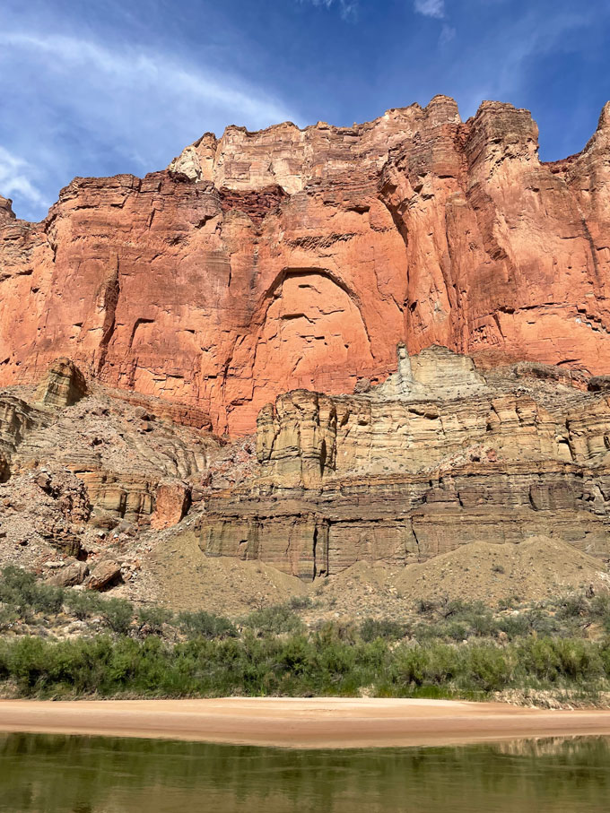 Colorful cliffs are shown rising over a sandy beach and river.