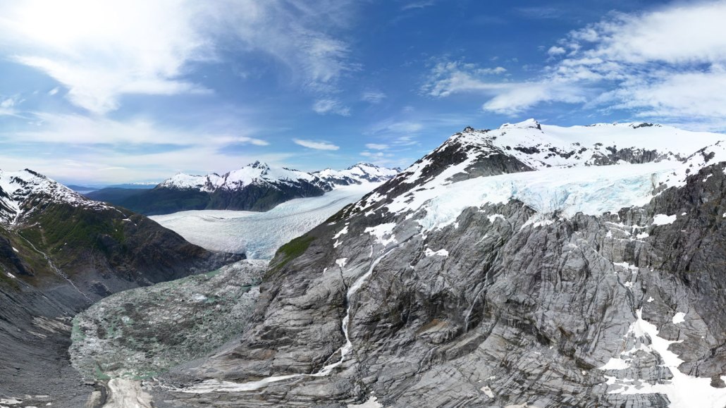 A panoramic view of a glacier nestled between snow-capped mountains under a partly cloudy blue sky. The glacier’s ice flows down into a rocky valley, with areas of broken, dirty ice near the base.