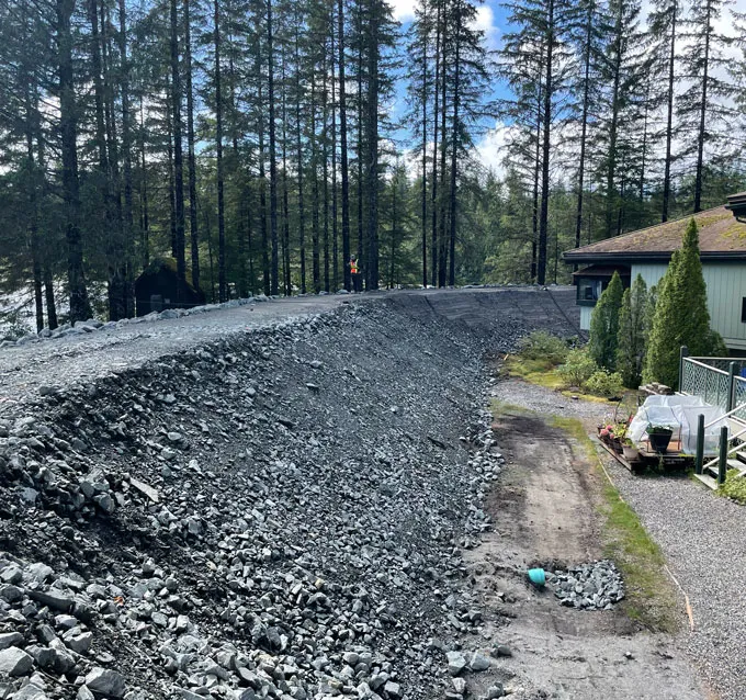 A sloped embankment of loose gray gravel rises beside a house. The embankment runs into a dense forest of tall evergreen trees. A person in an orange safety vest stands in the distance on the road.