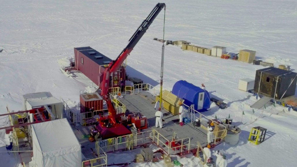 A research team in white protective suits works on a platform in Antarctica with a red crane lowering equipment into the ice. Portable shelters and supply containers surround the site.