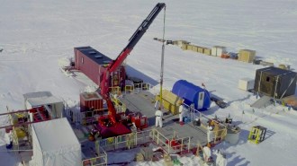 A research team in white protective suits works on a platform in Antarctica with a red crane lowering equipment into the ice. Portable shelters and supply containers surround the site.